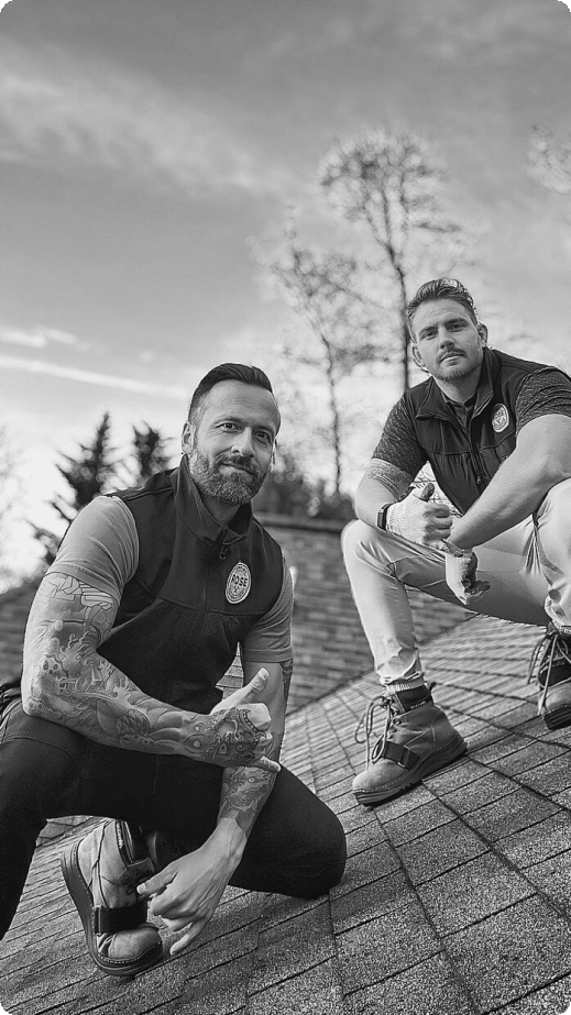 Two men squatting on a cobblestone path, one with a beard and sleeve tattoos, both wearing vests and looking at the camera. the background shows a cloudy sky and blurred trees. image in black and white.