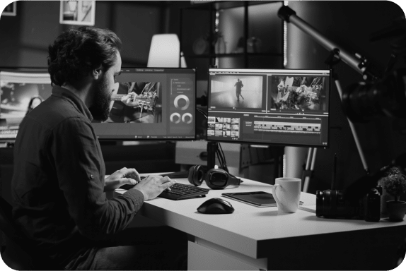 Black and white image of a man at a desk working on video editing. He is using a keyboard and mouse to operate multiple monitors displaying various editing software and footage. The workspace includes a camera, headphones, a cup, and other equipment.