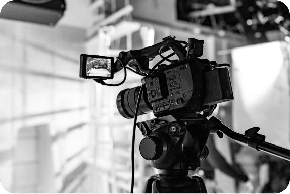 A black and white image of a professional video camera mounted on a tripod, with its external monitor displaying the scene it is recording. The background appears to be a studio setup with lighting equipment and other production gear visible.