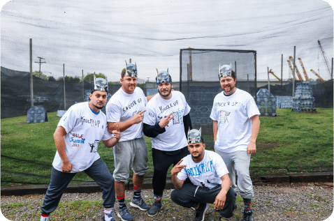 A group of five men wearing Batman-themed paper hats and white T-shirts poses outdoors in front of a paintball field. They seem to be in a playful mood, striking various poses, with one kneeling in front. The grassy field and paintball structures are visible behind them.