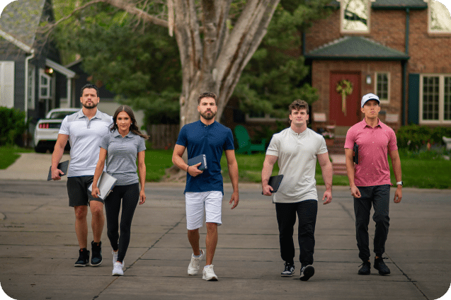 Five people, three men and two women, walk confidently down a suburban street holding laptops and notebooks. They are dressed in casual to sporty attire and appear focused. The background shows houses, trees, and parked cars.