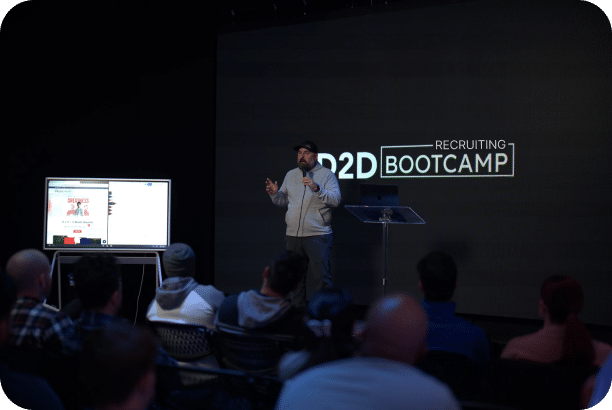 A person wearing a cap and hoodie speaks into a microphone on a stage in front of a large screen displaying "D2D Recruiting Bootcamp." An audience is seated and watching, and a smaller screen next to the speaker shows additional content.