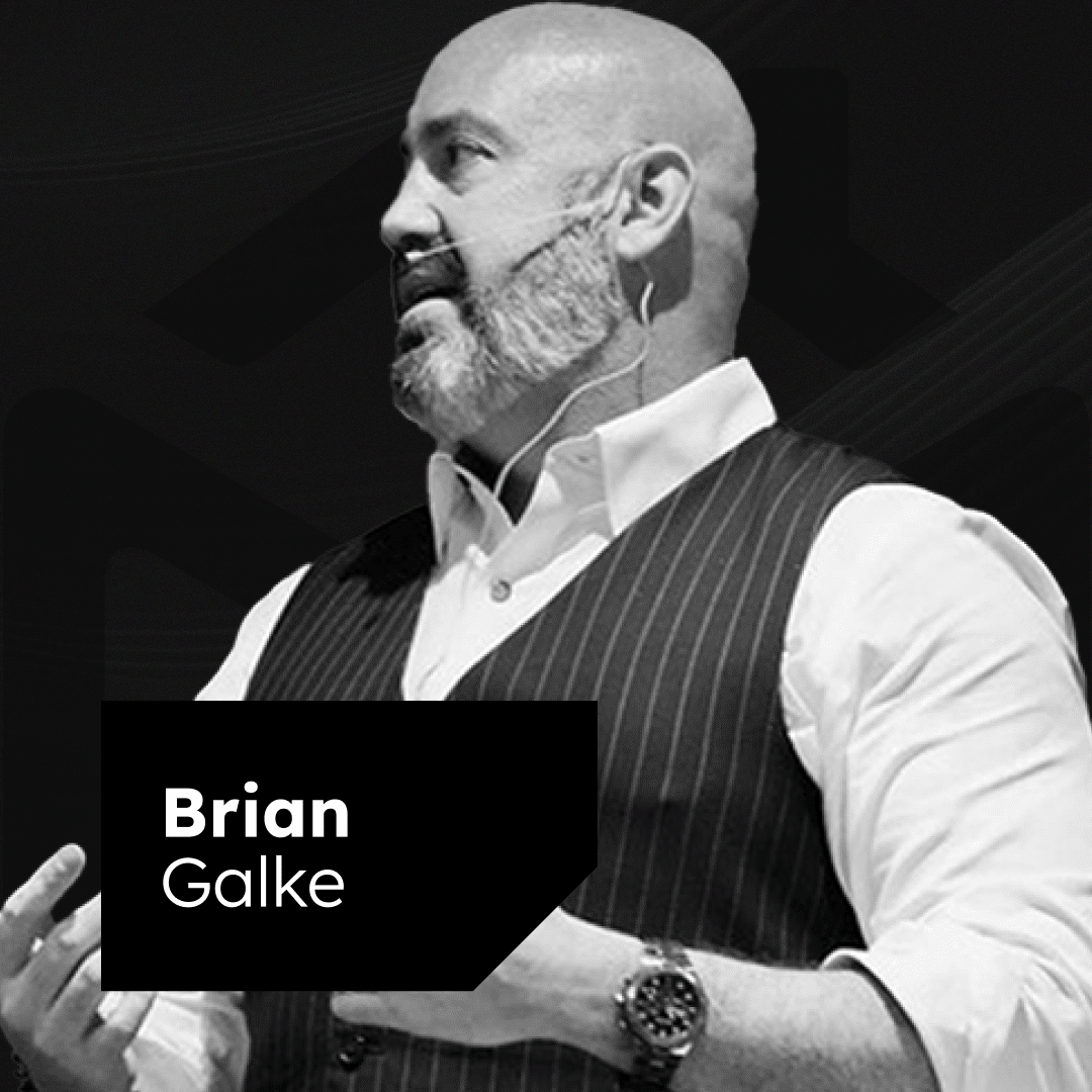 A bald man with a beard is speaking, wearing a white shirt and a dark pinstriped vest. He has a microphone headset and gestures with his hands. A text box on the image reads "Brian Galke." The background is dark and abstract.