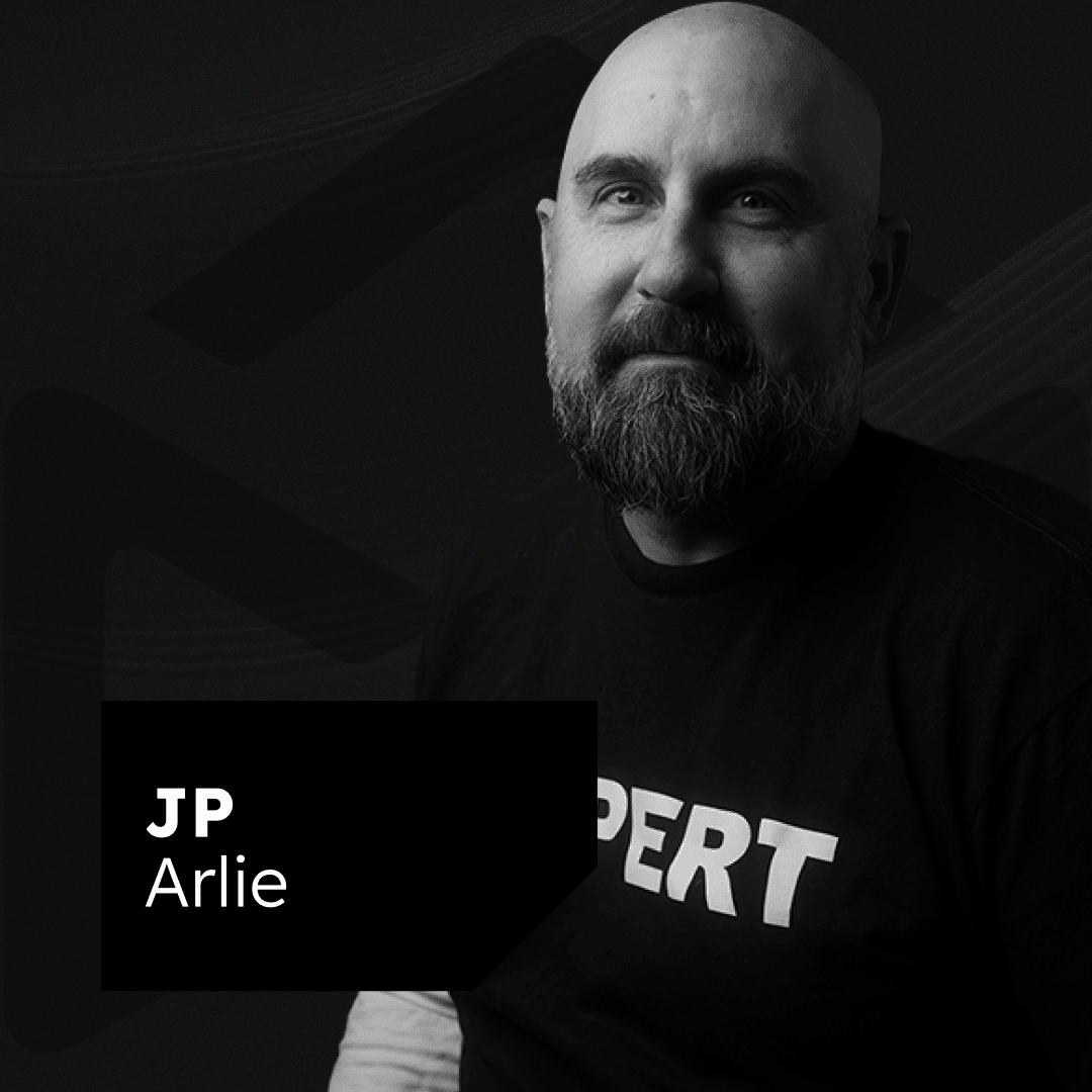 Black and white image of a bearded man with a bald head, wearing a shirt with the text "EXPERT" on it. He is looking directly at the camera. The text overlay at the bottom left corner reads "JP Arlie.