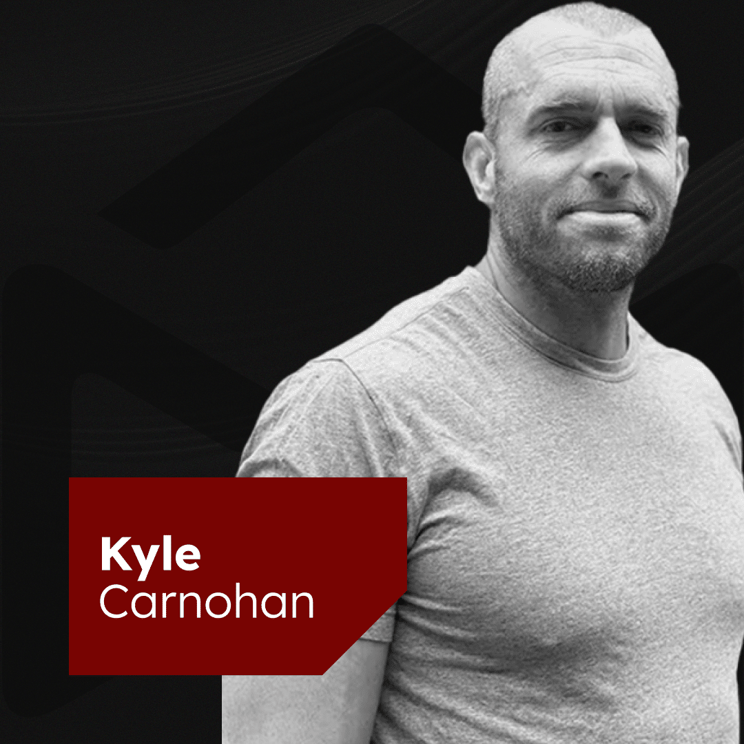 Black and white image of a man with a shaved head and a short beard wearing a plain T-shirt. He is looking at the camera with a neutral expression. A red rectangular banner in the bottom left corner contains the name "Kyle Carnohan" in white text.