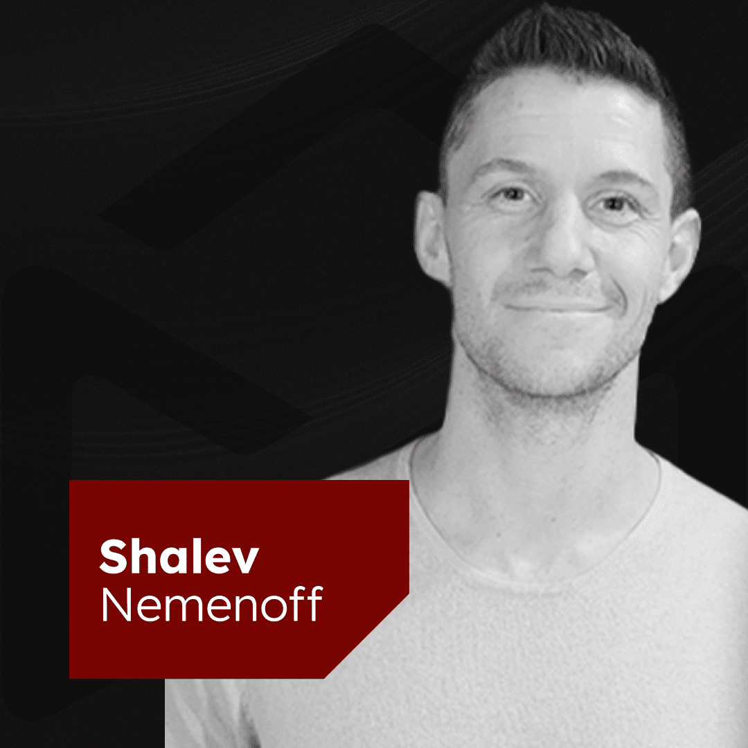 A grayscale image of a person smiling while facing the camera. They are wearing a light-colored shirt. There is a red rectangle at the bottom left corner with the text "Shalev Nemenoff" written in white. The background is dark with subtle patterns.