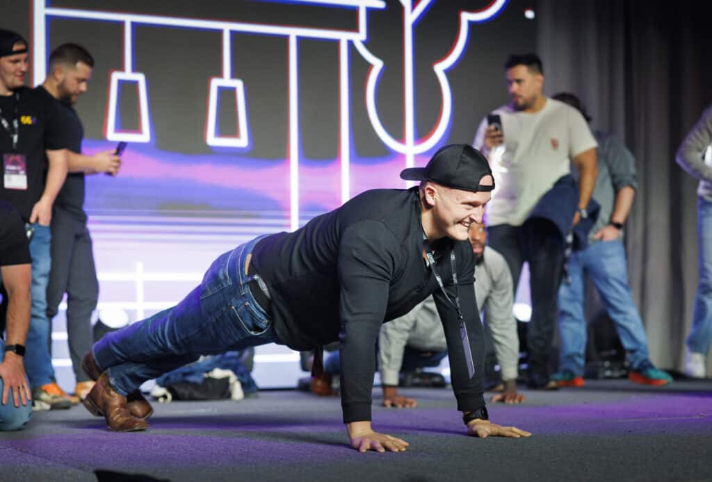 A man in a black shirt and jeans performs push-ups on stage while smiling. He wears a backwards cap. Several onlookers in casual clothing watch and take photos, standing on a stage with colorful lighting and abstract background designs.