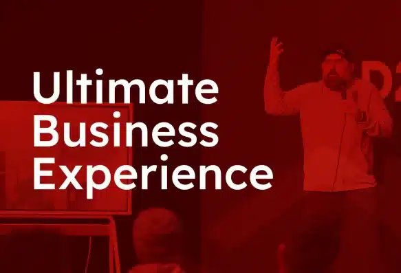 A speaker energetically presents on stage. The background is a red overlay with the text "Ultimate Business Experience" in bold white letters. Audience members are visible in the foreground.