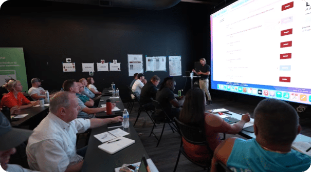 A group of people sitting at tables in a dark room, attentively listening to a presentation. The presenter stands near a large, colorful screen displaying a website or application interface. Notebooks and water bottles are on the tables.