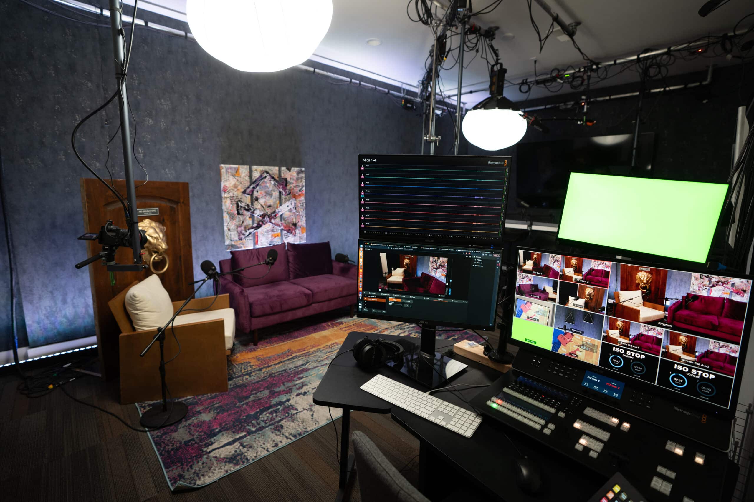 A multi-camera studio setup with two monitors displaying video feeds. A purple couch and a brown chair sit on a colorful rug. Overhead lights illuminate the room, and a green screen is visible in the background. Audio and video equipment are arranged on a desk.