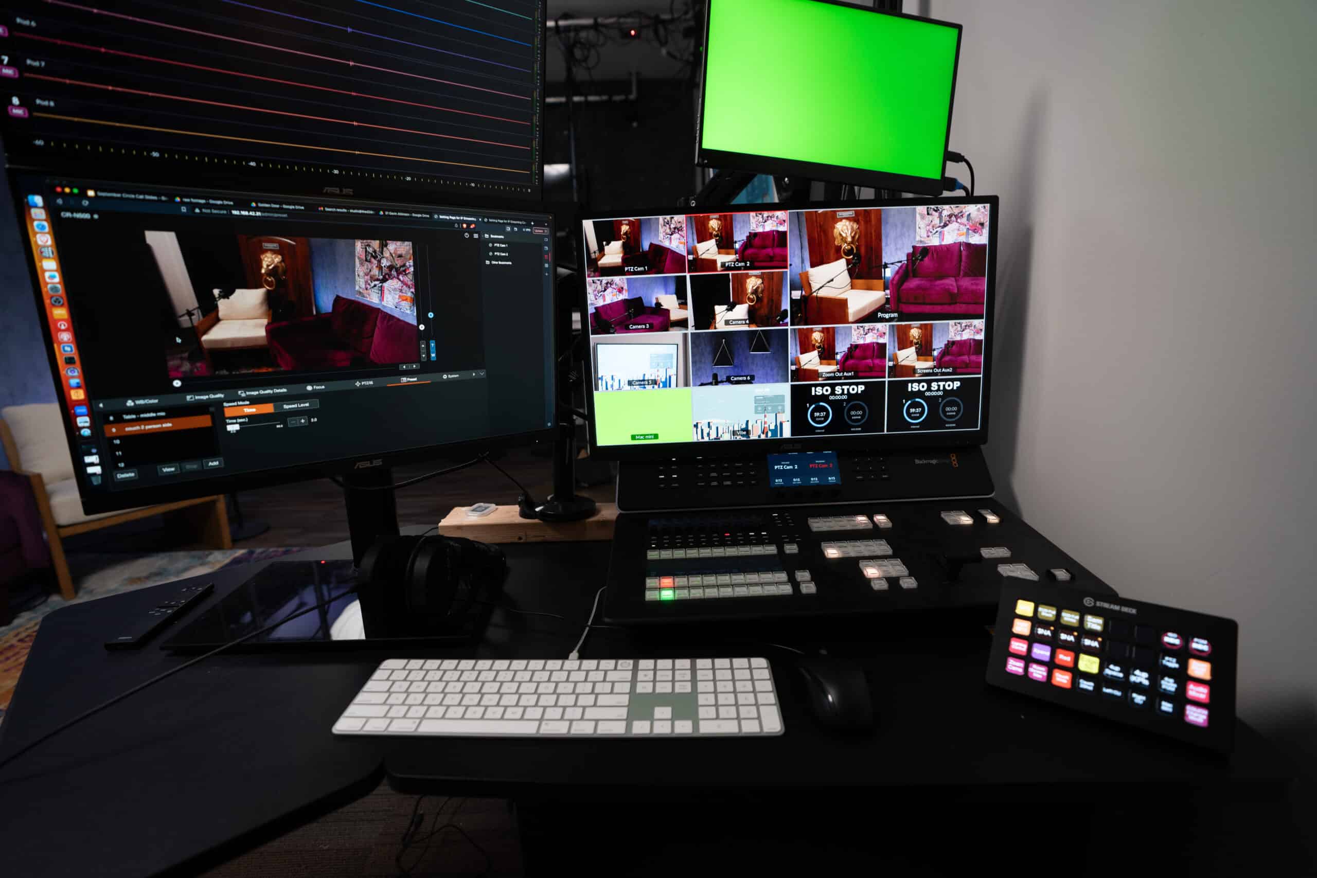A control room setup with multiple screens displaying video feeds. One monitor shows a grid of video thumbnails, another has editing software, and a small green screen is visible. Computer peripherals and a video switcher are on the desk.