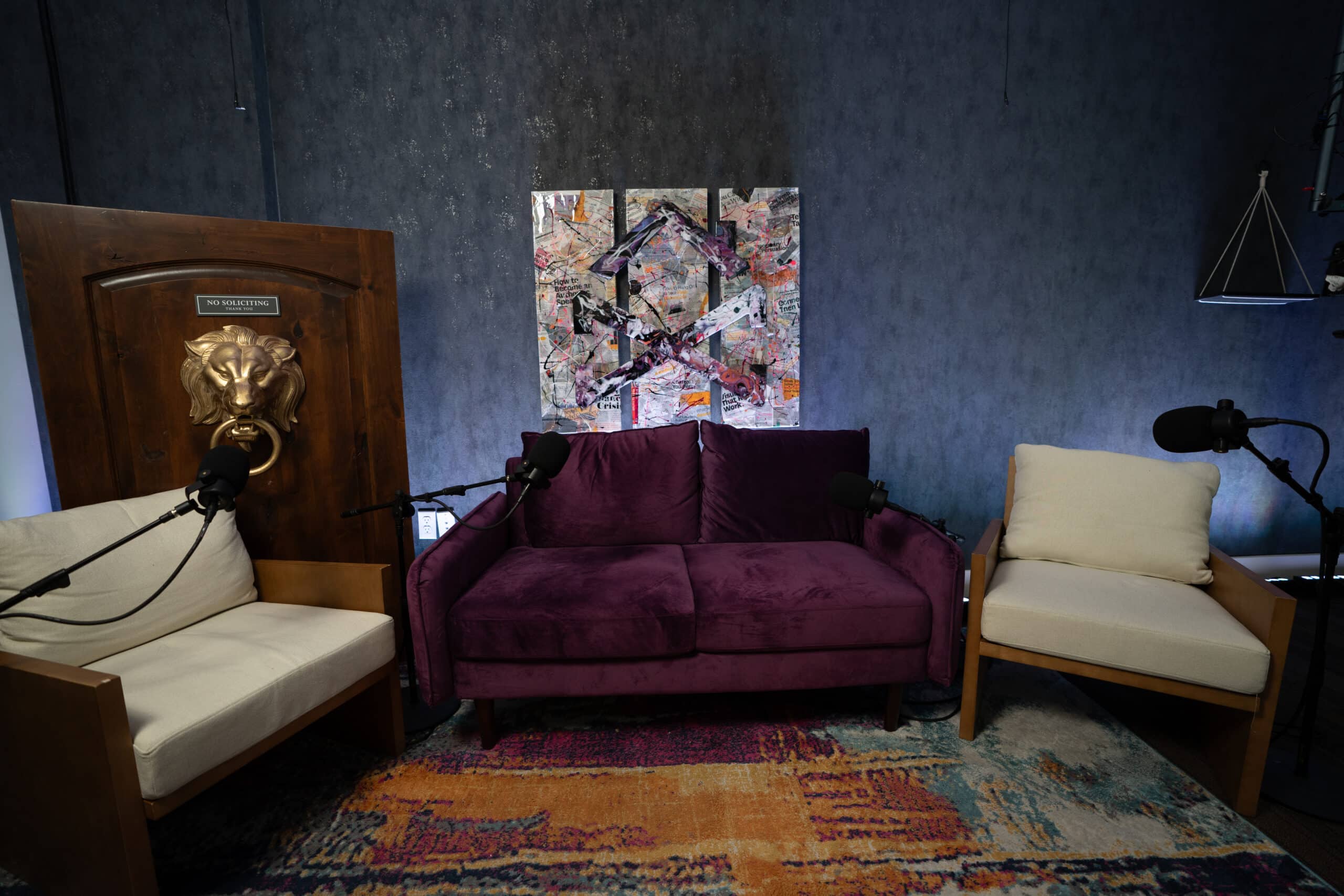 A cozy podcast studio setup with a purple sofa in the center, flanked by two armchairs. Microphones are positioned in front of each seat. Behind the sofa, a colorful abstract artwork is displayed on a dark textured wall.