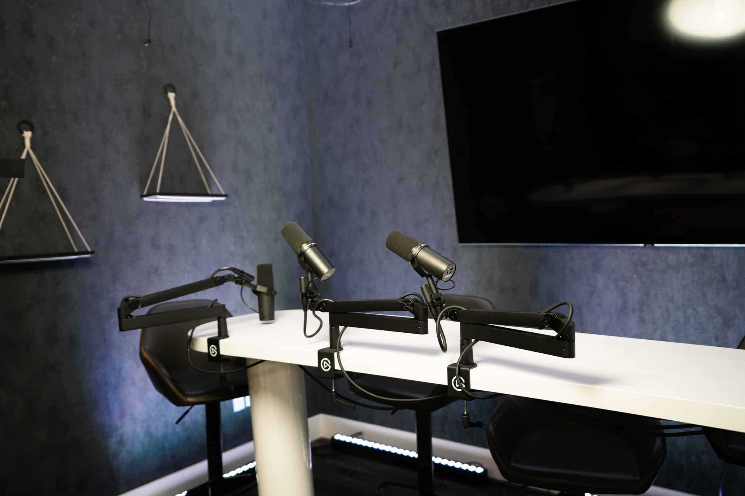 A modern podcast studio with a white table and two black microphone arms. Three black chairs are arranged around the table. A large flat-screen TV is mounted on the wall, and the room is dimly lit with sleek decor.