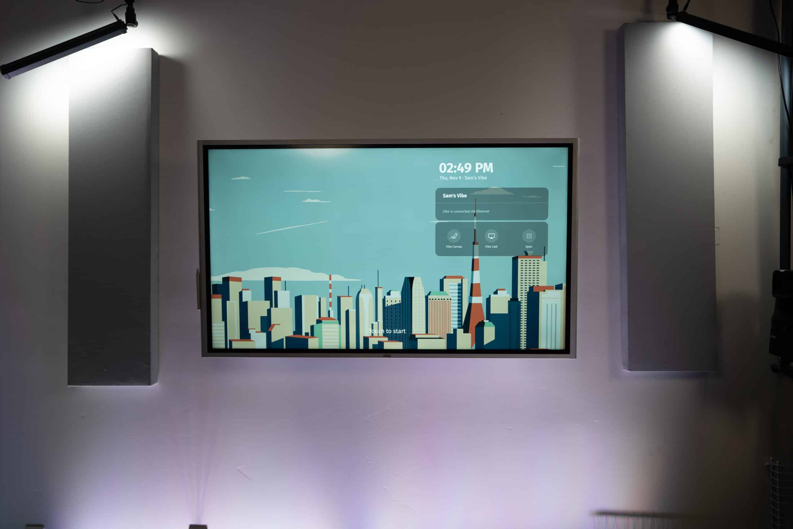 A digital screen displaying the time 02:49 PM and a weather forecast is mounted on a wall. The screen shows a stylized cityscape with skyscrapers. Two wall-mounted lights illuminate the space.