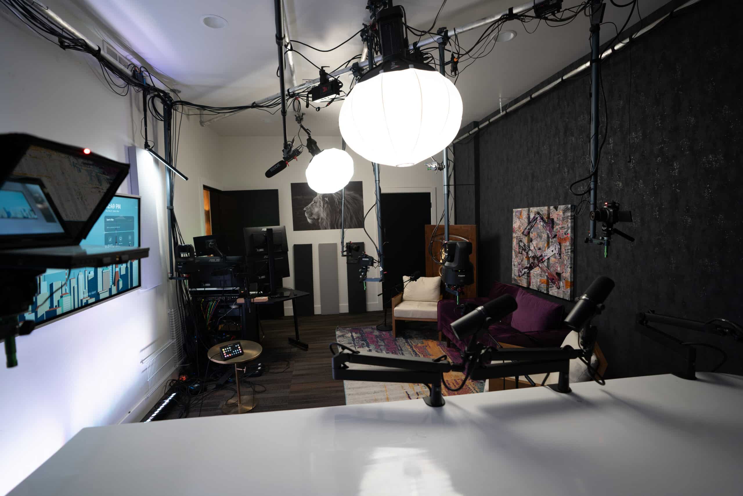 A modern recording studio with various cameras, microphones, and lighting equipment. There's a bright white table in the foreground, a purple couch, and colorful artwork against a textured black wall in the background.