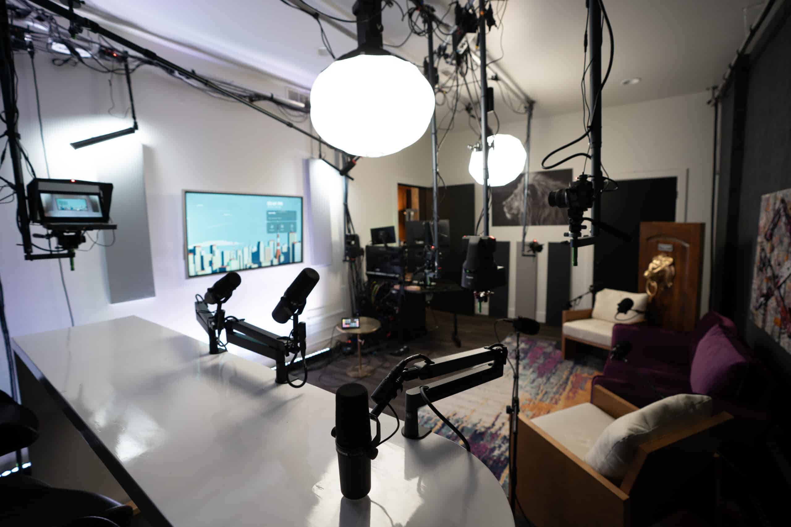 A podcast studio with multiple microphones on a table, bright overhead lights, and cameras positioned around the room. A TV screen displays graphics in the background, and there are couches and colorful rugs on the floor.
