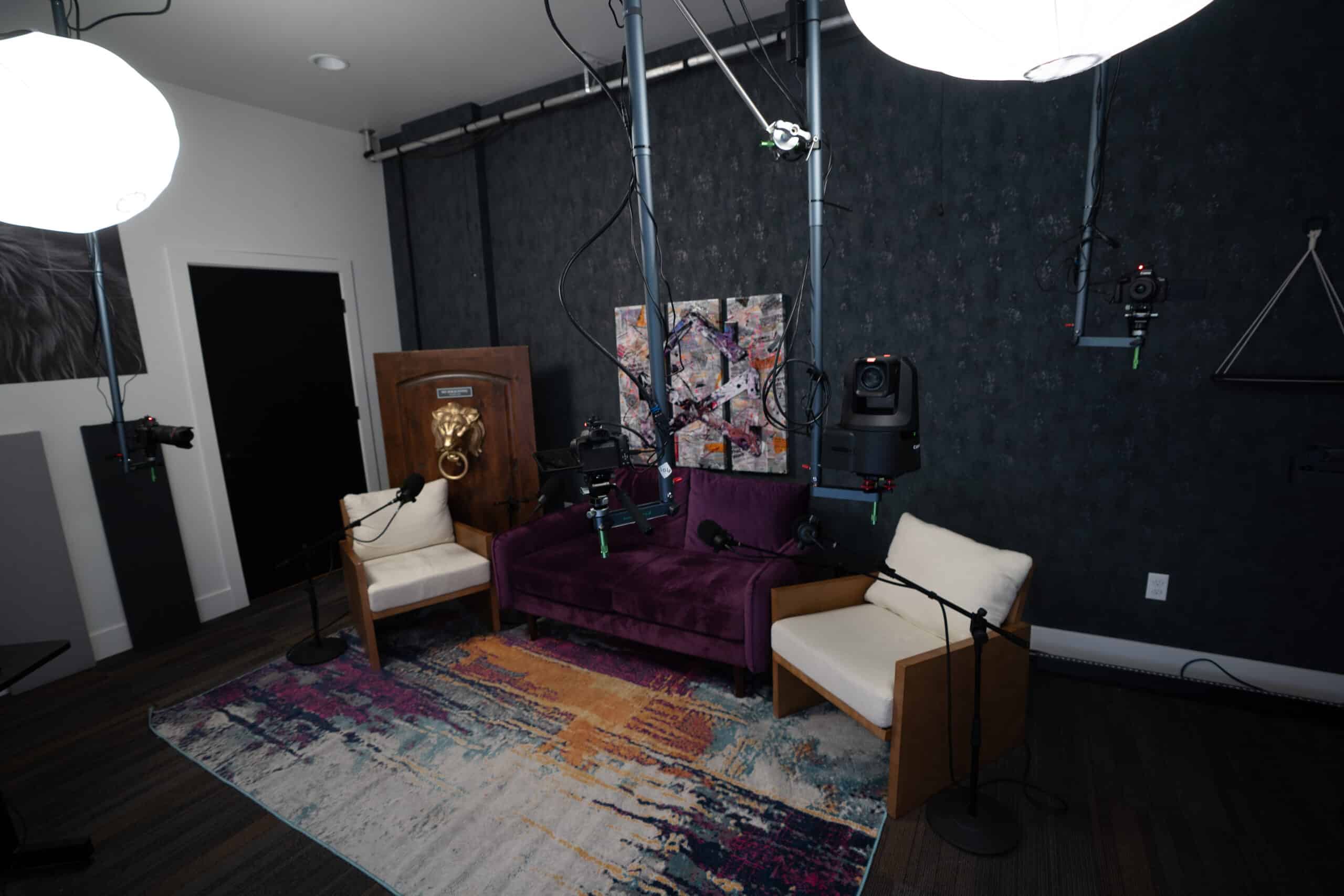 A modern podcast recording studio with a purple sofa, two white chairs, multiple microphones on stands, and cameras. The room features dark walls, a colorful rug, a lion head on the wall, and abstract art.