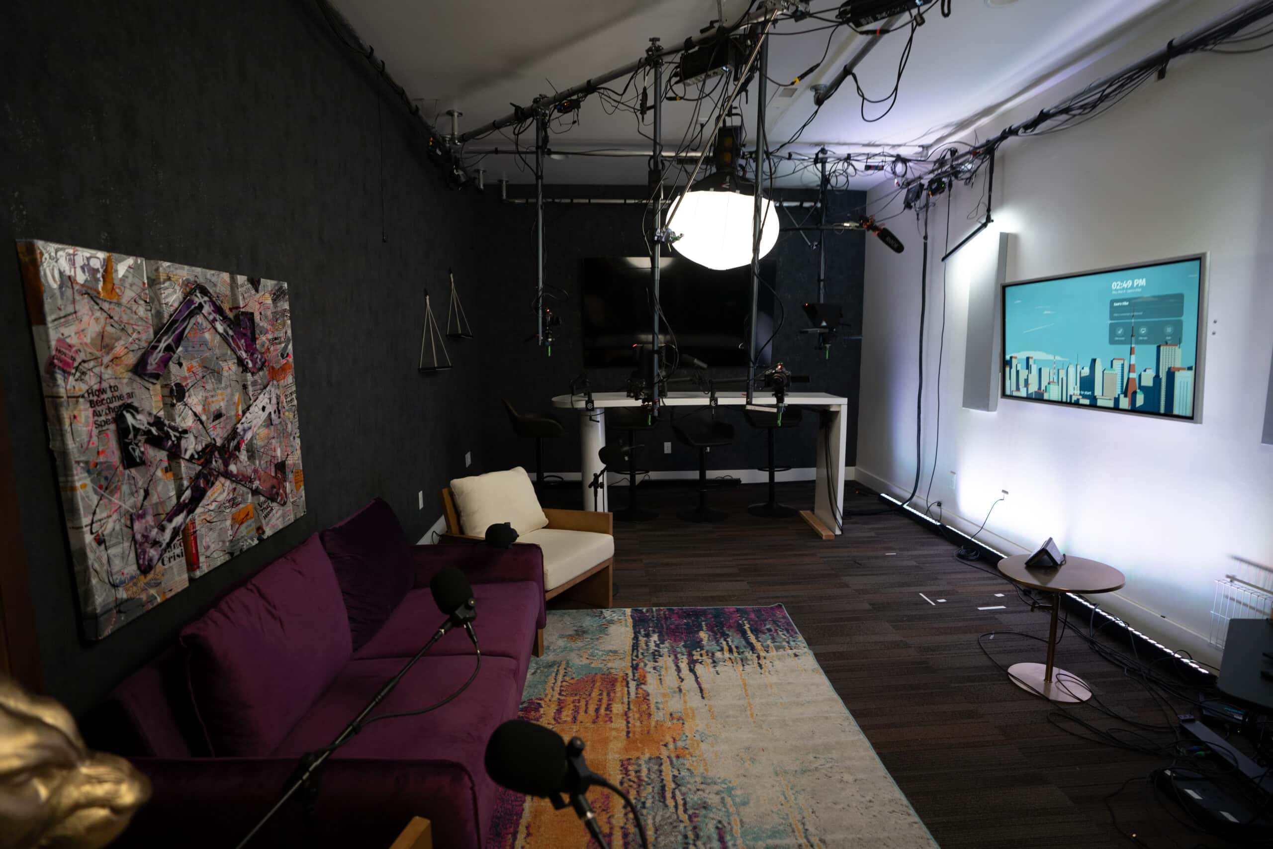 A dimly lit podcast studio featuring a dark wall with abstract art, a purple couch with microphones, and a colorful rug. Overhead lighting equipment is visible. In the background, there's a desk with chairs and a large screen on the wall.