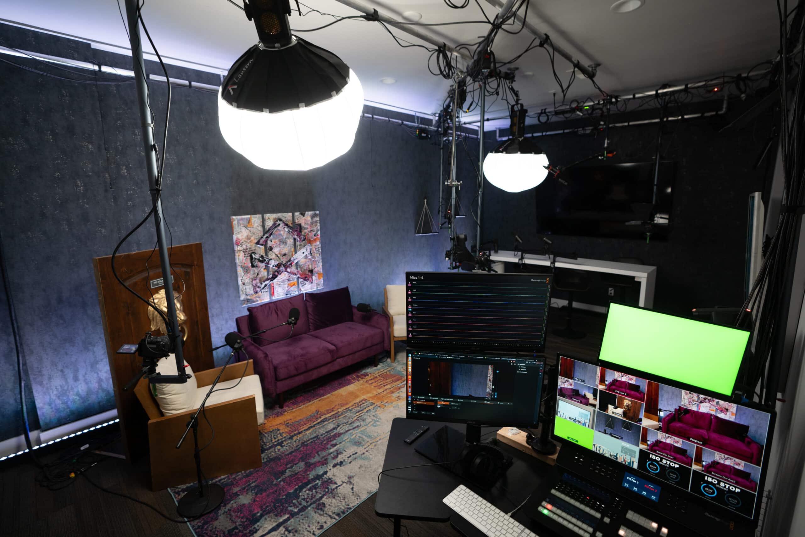 A small studio setup with cameras, lights, and monitors. A purple sofa and colorful rug are in the background, with a wall-mounted screen and cables running overhead. The control desk has multiple screens displaying a video editing interface.