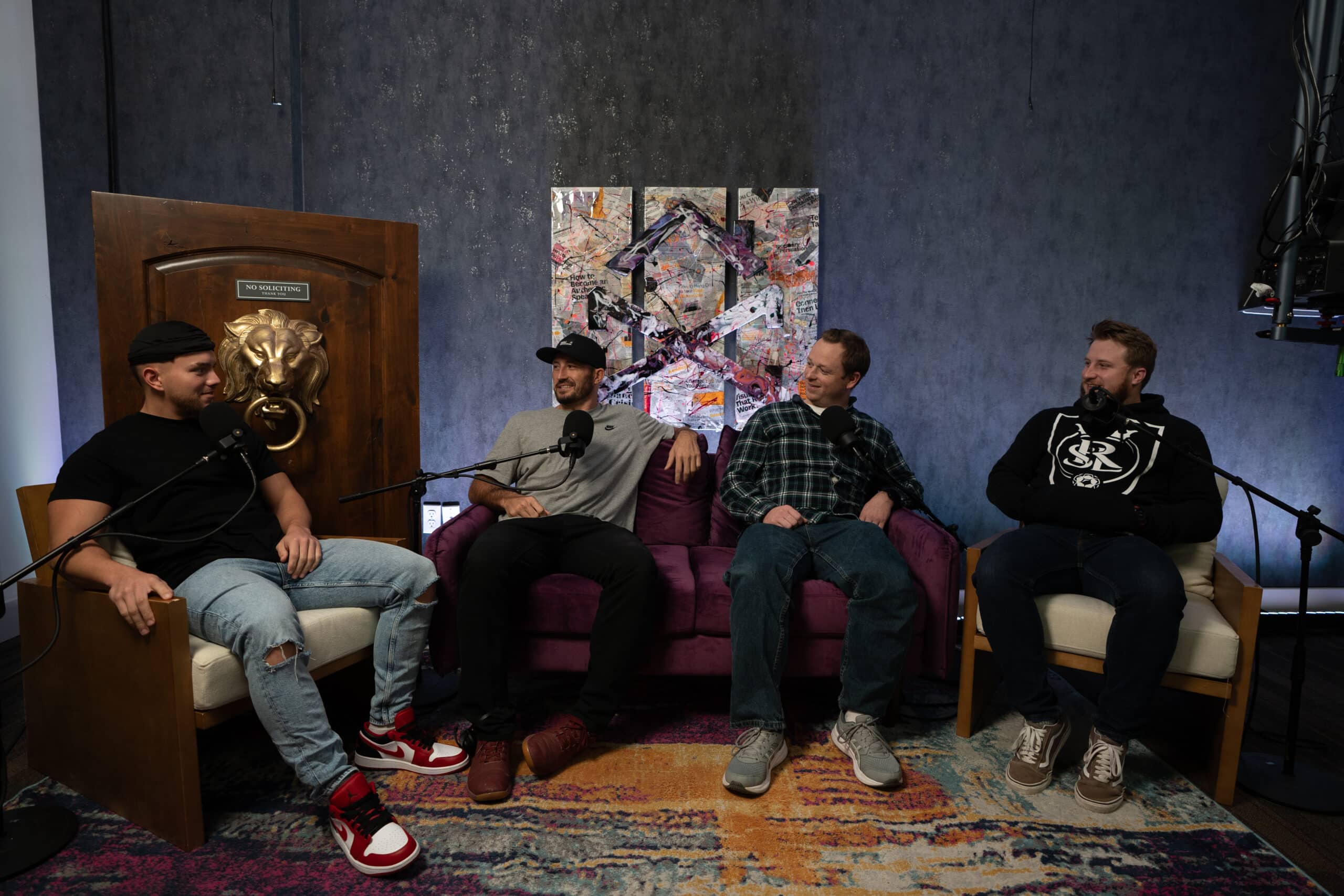 Four men are seated on a purple sofa and chairs, recording a podcast with microphones in a cozy room. The background features a large wooden door with a lion's head knocker and abstract art. A colorful rug is on the floor.