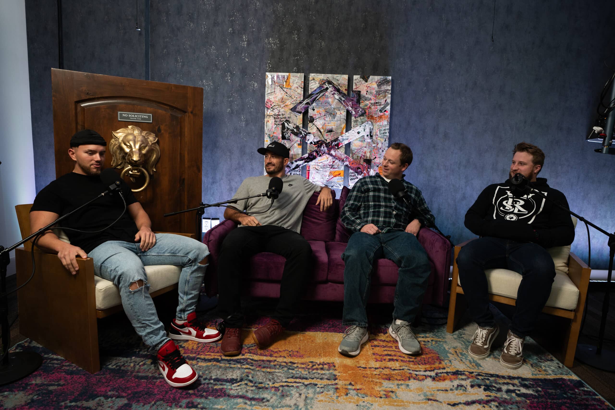 Four men are seated on a couch in a podcast setup with microphones. The background features a dark textured wall and eclectic artwork. A door with a large lion's head knob is on the left. The floor has a colorful rug.