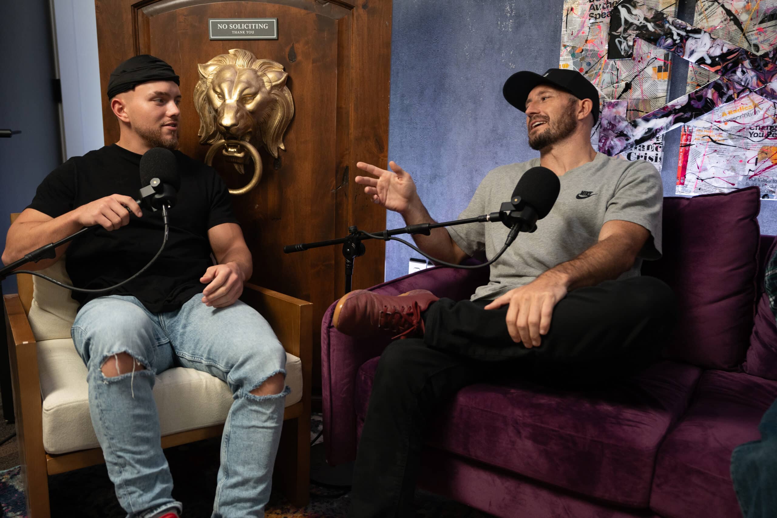 Two men sit in chairs, holding microphones, engaged in conversation. The backdrop features a wooden panel with a lion head decoration and a sign saying 