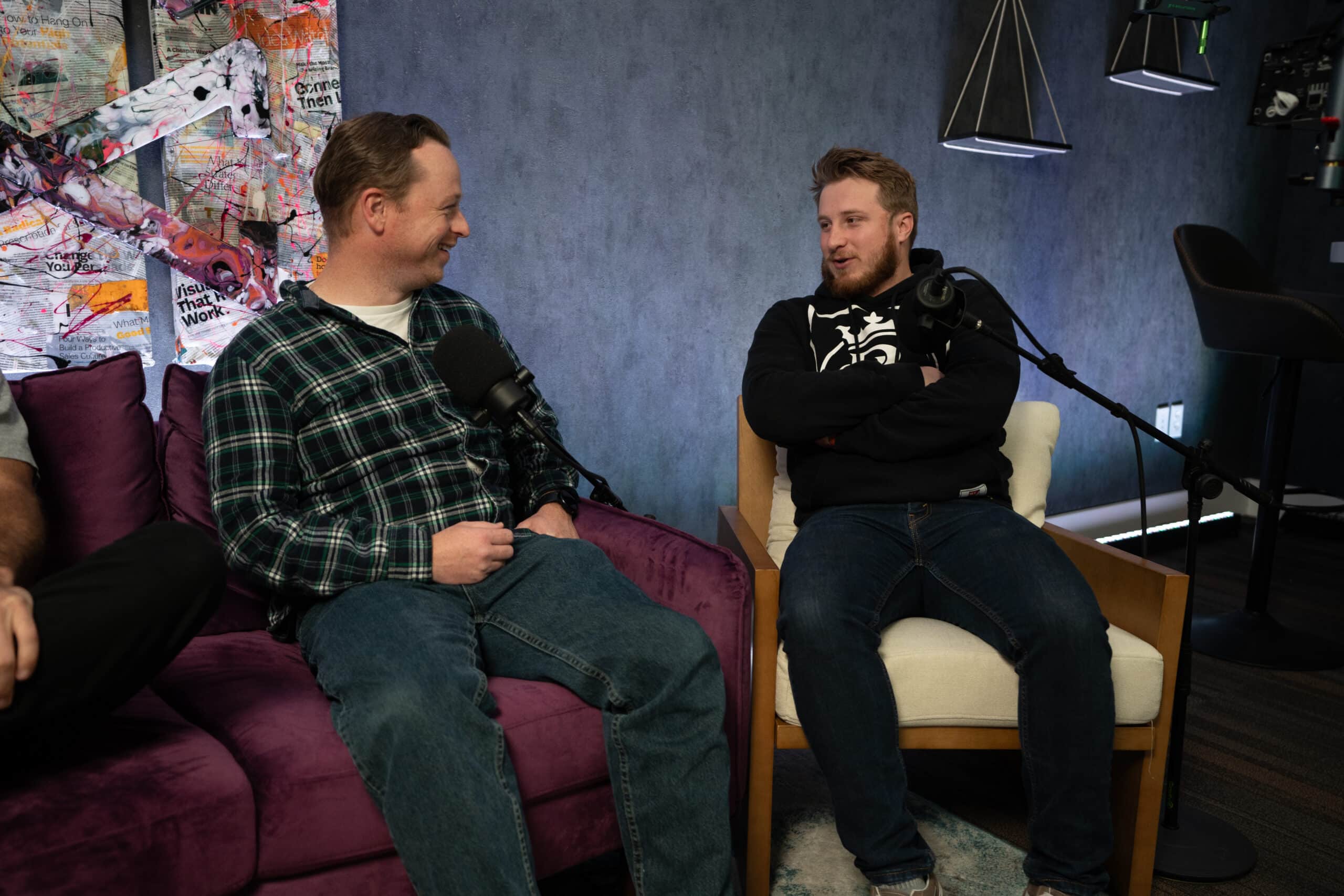 Two men sit in chairs facing each other, engaged in conversation, with microphones in front of them. One is on a purple couch, wearing a plaid shirt and jeans. The other is on a white armchair, wearing a black hoodie. The background has colorful art.