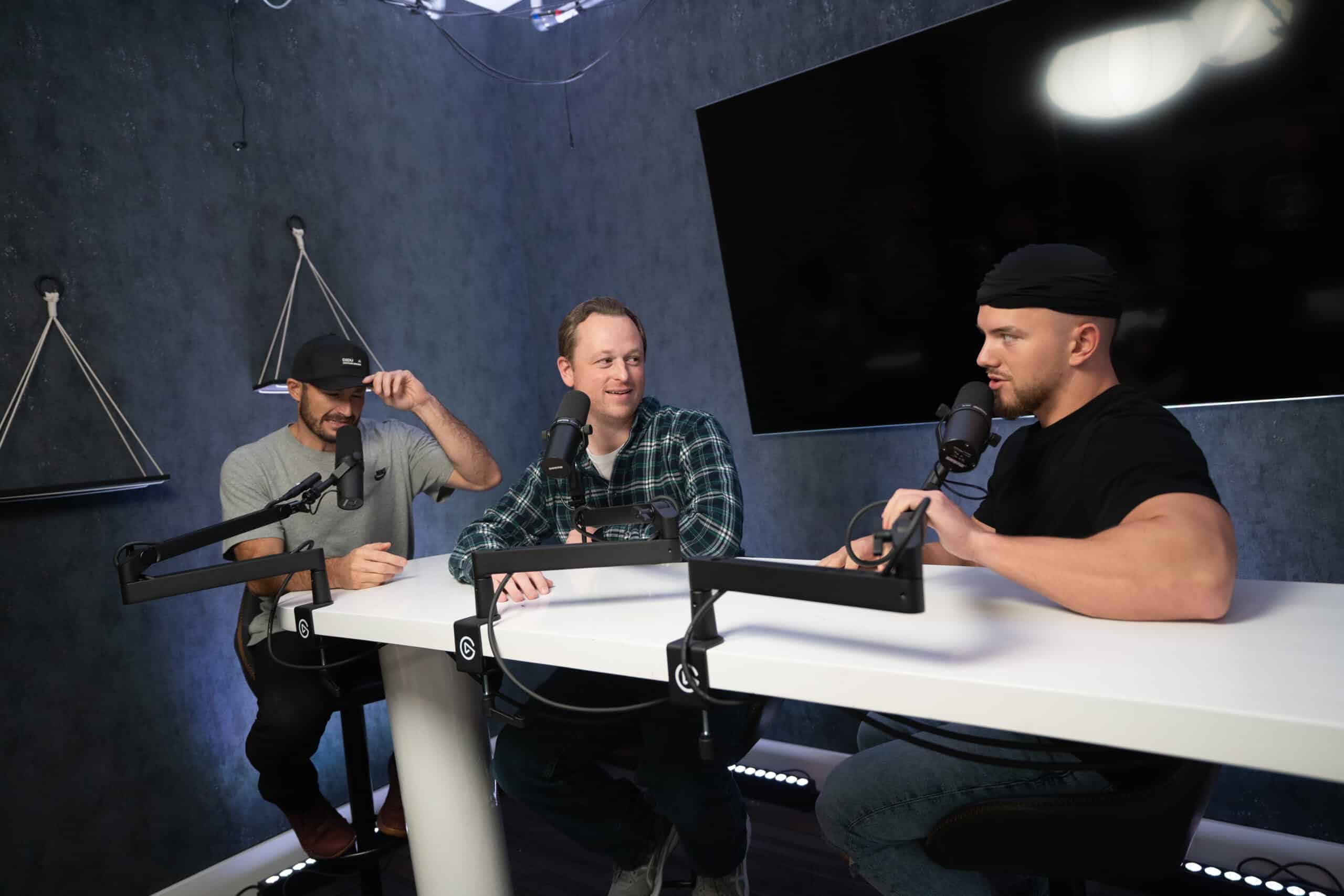 Three men are sitting at a table with microphones, engaged in conversation. The setting appears to be a podcast studio with a dark wall and a mounted flat-screen TV in the background. The men are casually dressed and appear animated in discussion.