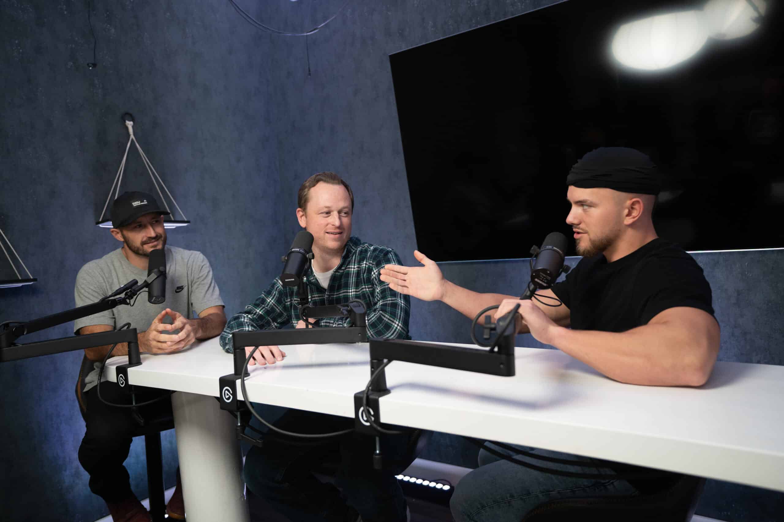 Three men sit at a table with microphones in front of a large screen in a podcast studio. One man in a cap gestures while speaking, another wears a plaid shirt, and the third listens attentively. The room has a modern, tech-friendly ambiance.
