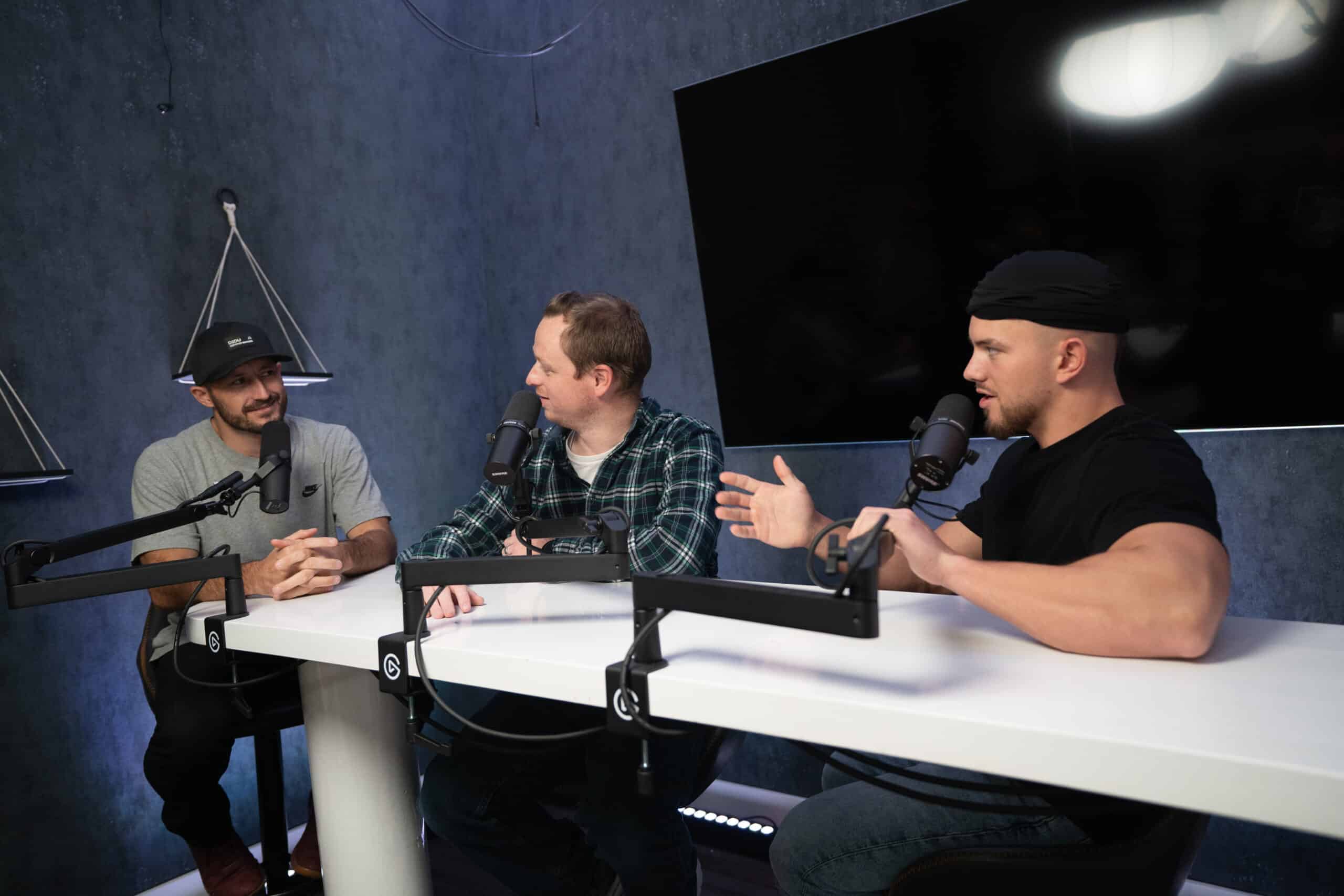 Three men are sitting at a table with microphones, engaged in a conversation. The two on the ends are wearing hats, while the man in the middle is in a plaid shirt. A large screen is mounted on the wall behind them in a dark, modern studio.