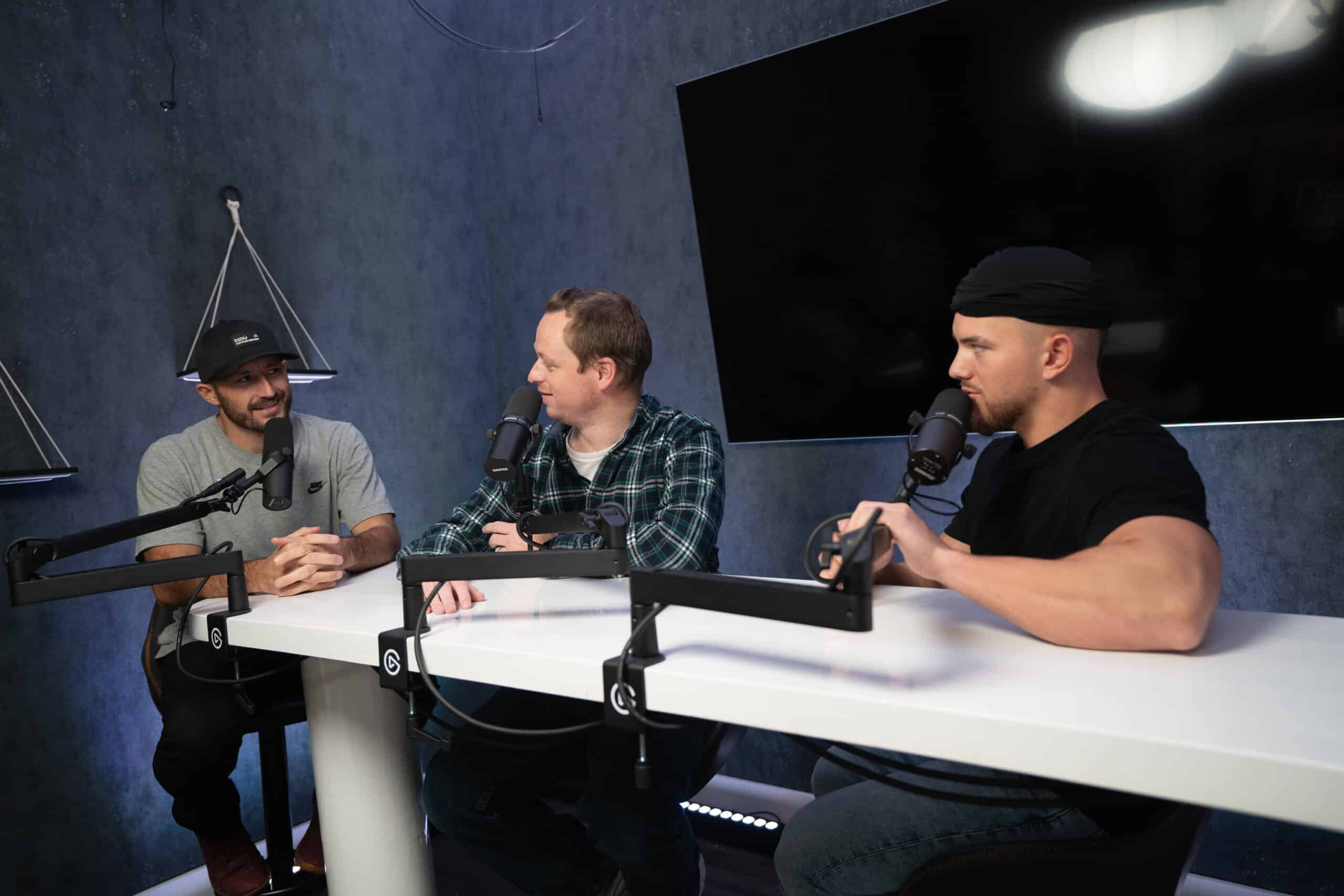 Three men sit at a white table with microphones, engaged in conversation. The room has dark walls and a circular light fixture overhead. Two men wear caps, and one wears a plaid shirt. They appear to be recording a podcast or radio show.