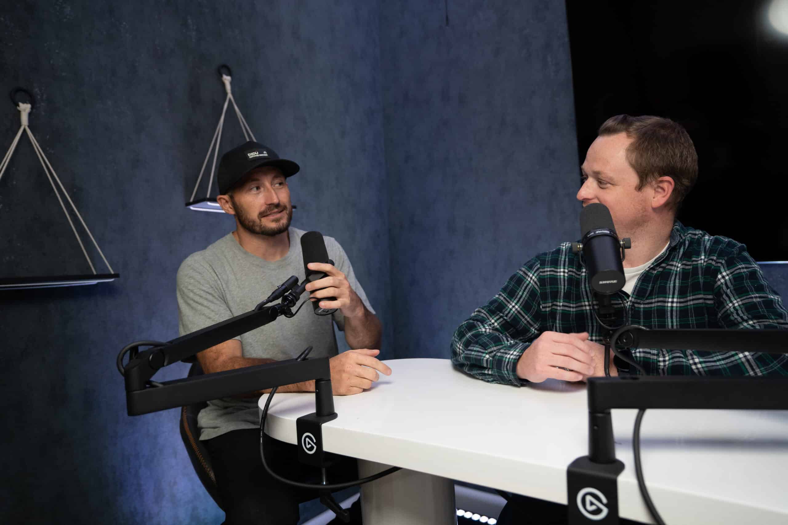 Two men are talking into microphones in a podcast studio. One wears a black baseball cap and gray T-shirt, the other wears a green plaid shirt. They are seated at a white table with podcast equipment. The background is dark with hanging wall decor.