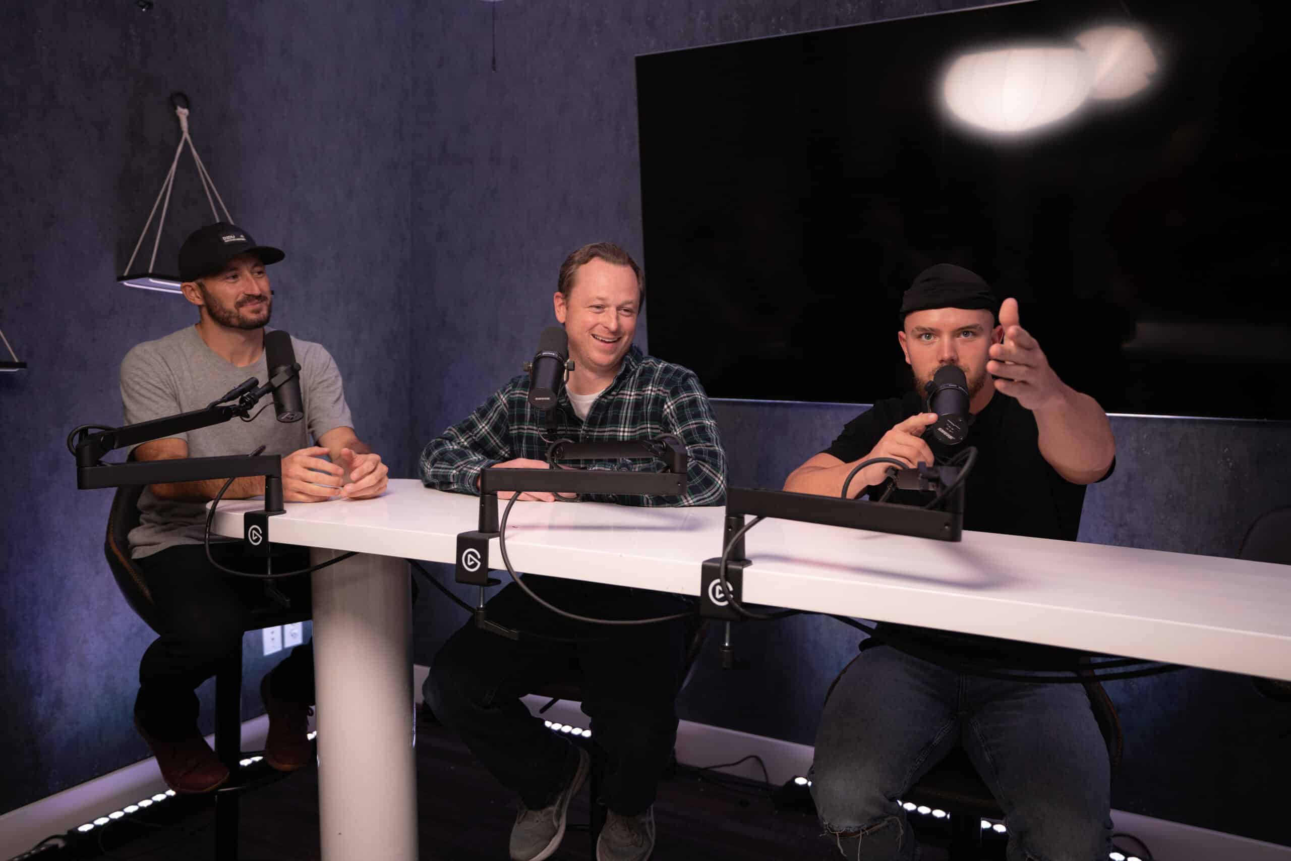 Three people are sitting at a table in a recording studio, each in front of a microphone. The person on the right gestures forward, while the one in the middle is smiling. A large screen is mounted on the wall behind them.