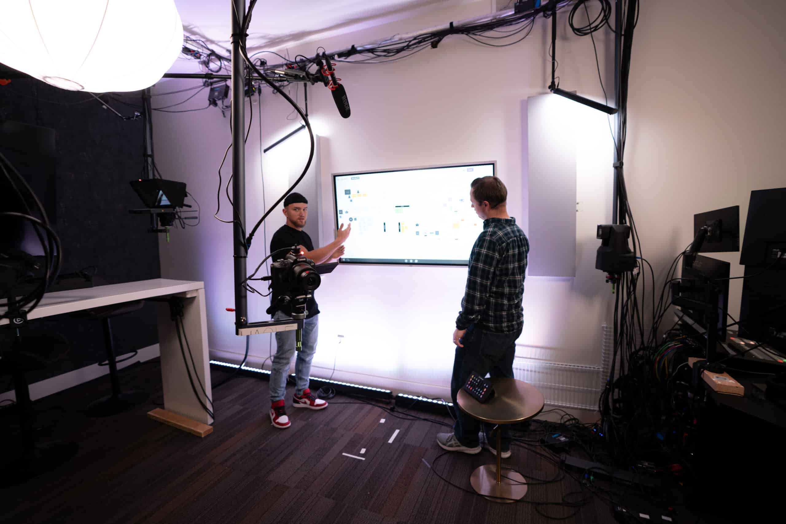 Two people in a studio setting are discussing in front of a large screen displaying a diagram. One person is gesturing at the screen, while the other listens. The room is equipped with camera and lighting gear.