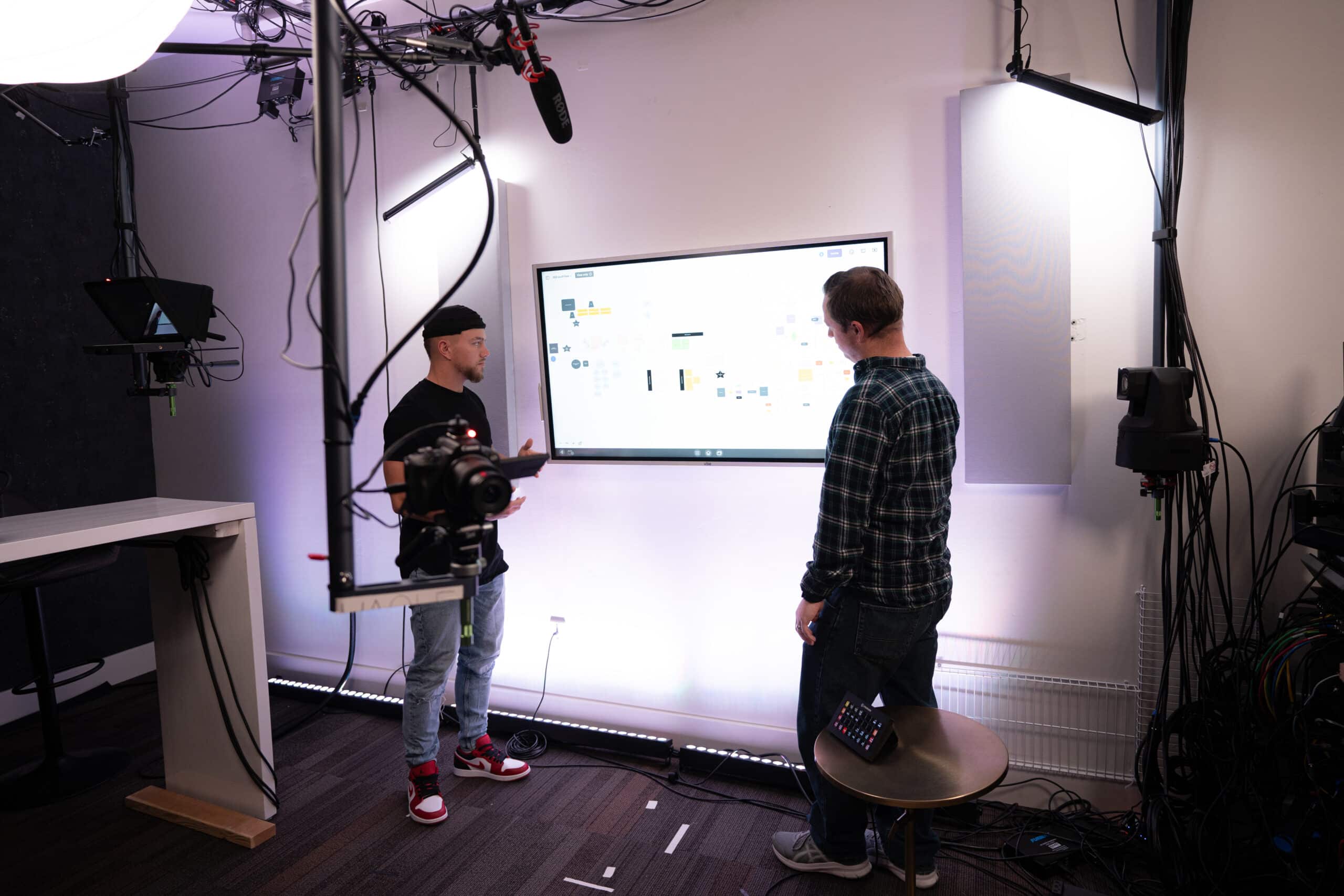 Two people stand in a room with studio lighting and equipment, looking at a large screen displaying various icons and diagrams. A camera is mounted nearby, along with wires and a microphone above.