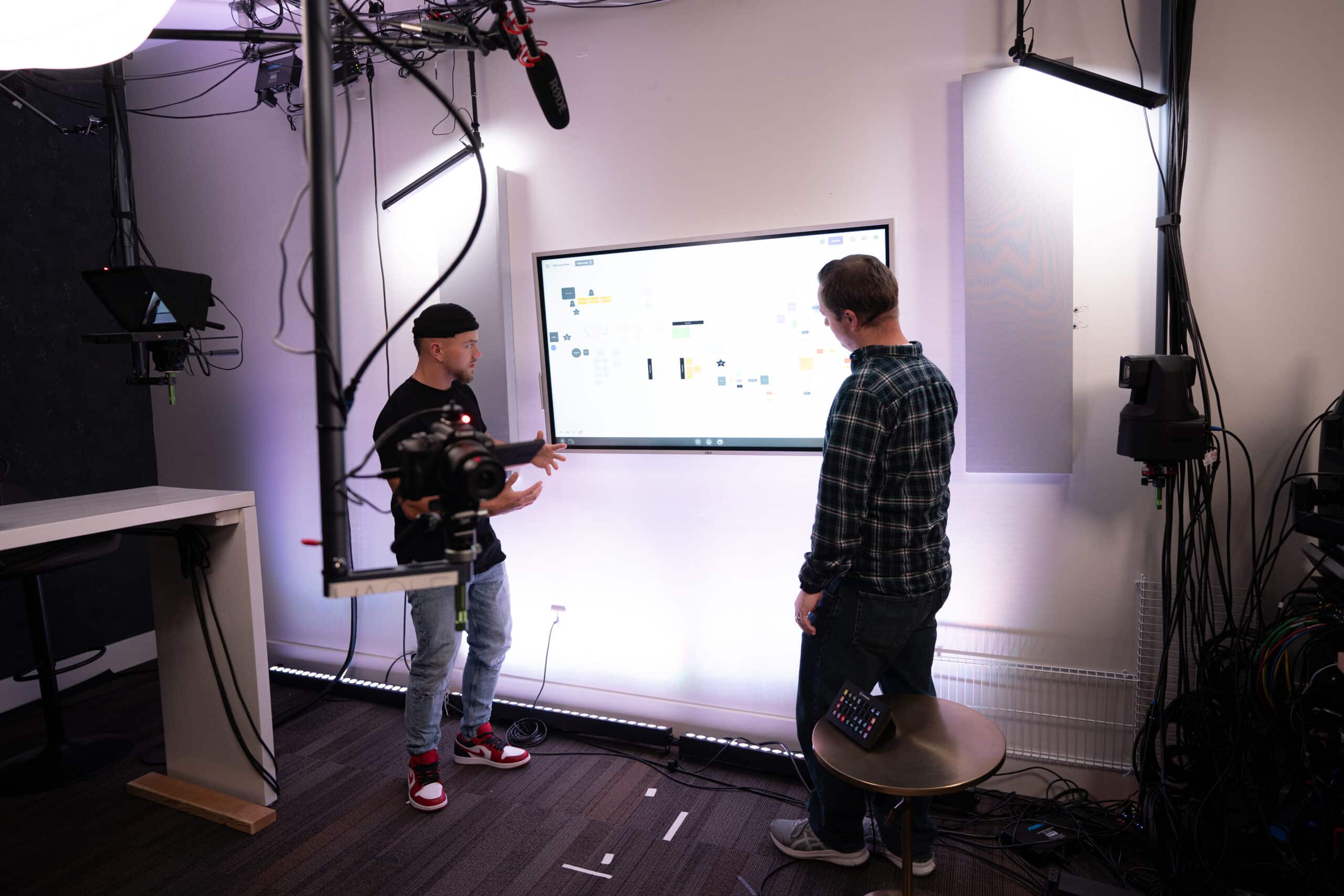 Two people are standing in a studio setting with cameras and lighting equipment. They are looking at a large screen displaying a diagram or flowchart. One person gestures toward the screen while the other observes.