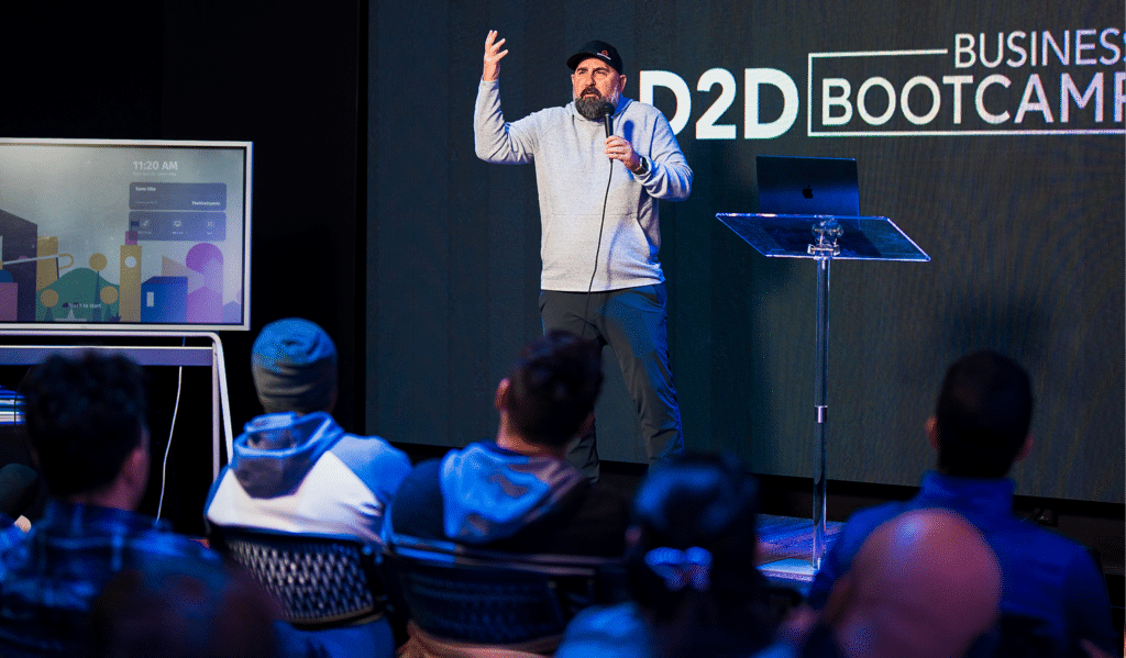 A man in a grey sweater and black cap speaks into a microphone on stage at a business bootcamp event. An audience listens, seated in front of him. A screen and a podium with a laptop are visible on stage.