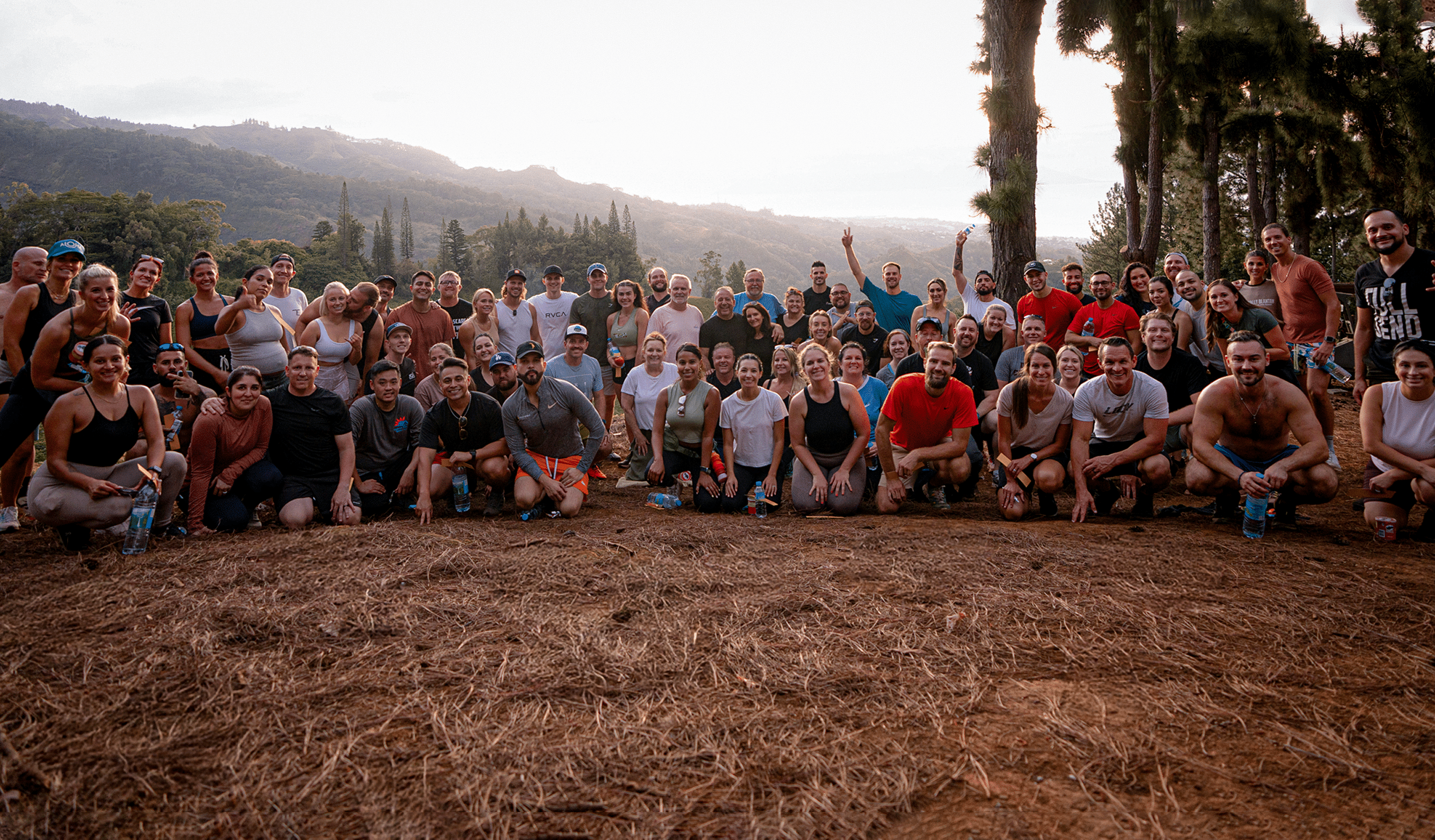 A large group of people are gathered outdoors on a forested hillside, smiling and posing for a photo. They appear to be participating in a fitness or outdoor event, surrounded by trees and mountains in the background.