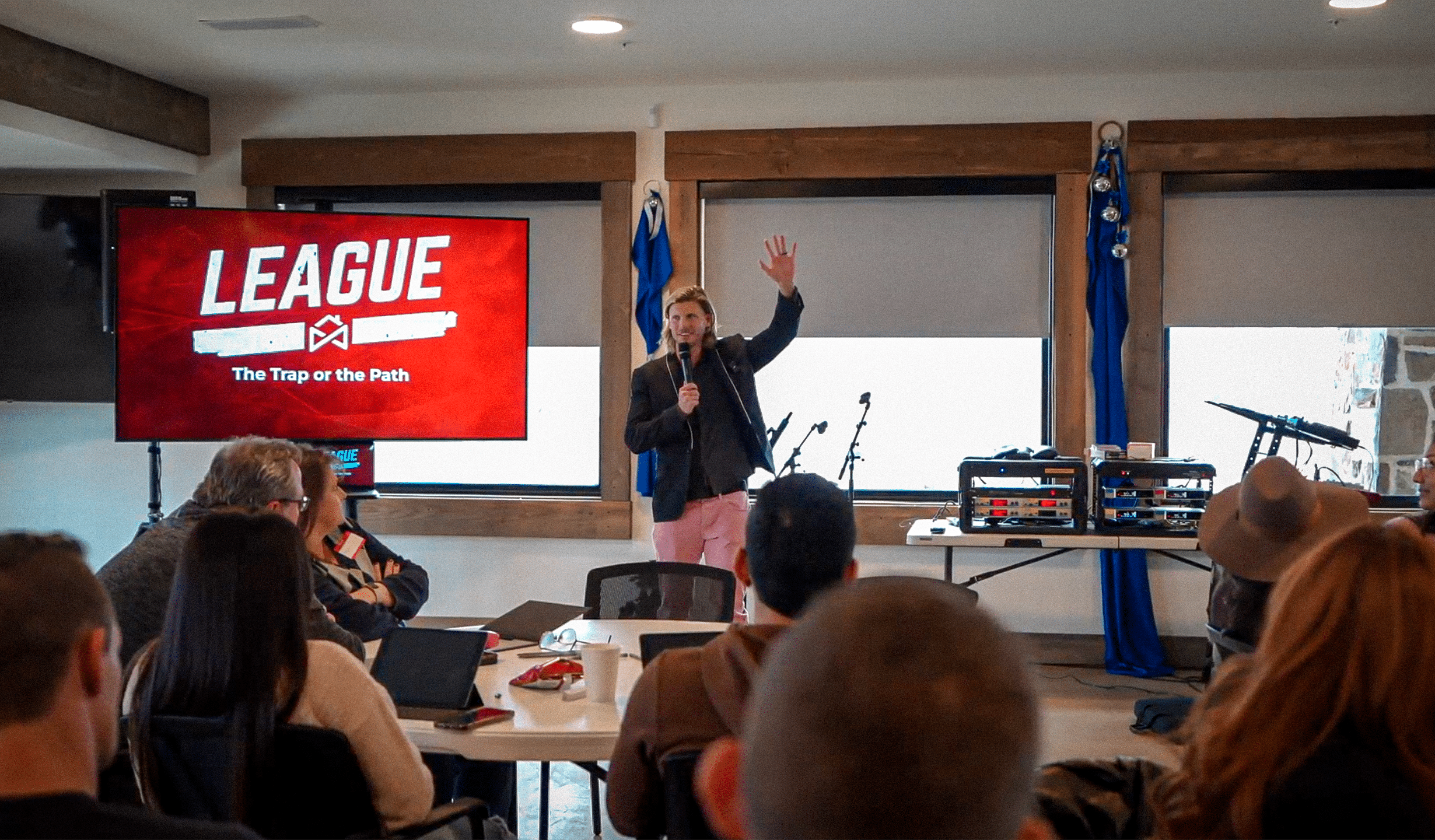 A person is speaking with a microphone in front of an audience in a room. A screen displays the title "LEAGUE The Trap or the Path." People are seated at tables with laptops and drinks, facing the speaker. Musical equipment is visible in the background.