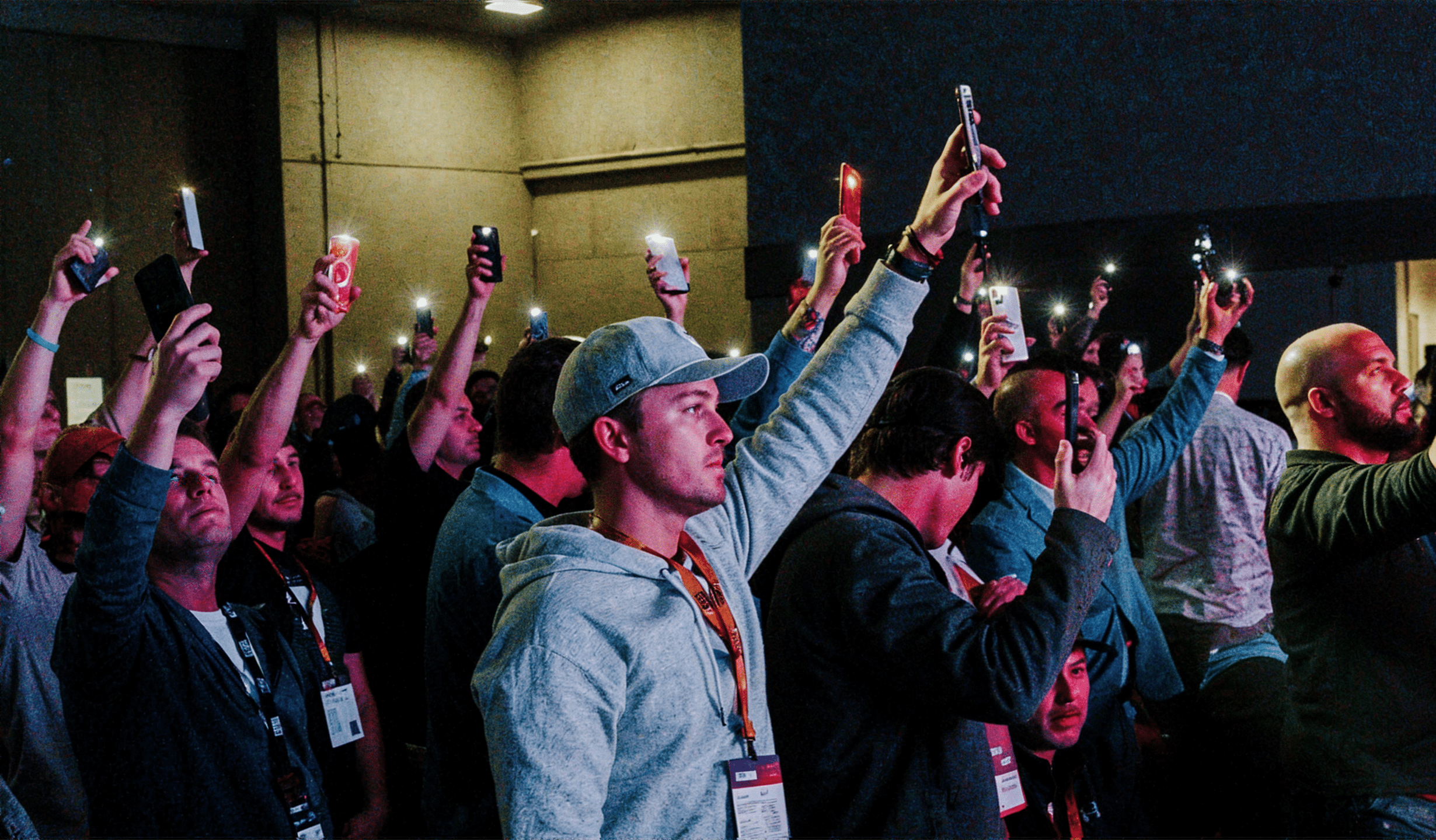 A crowd of people stands indoors, holding up lit smartphones in a darkened room. The individuals appear focused on an event or performance. Many are wearing casual clothing and event badges. The atmosphere is lively and engaged.