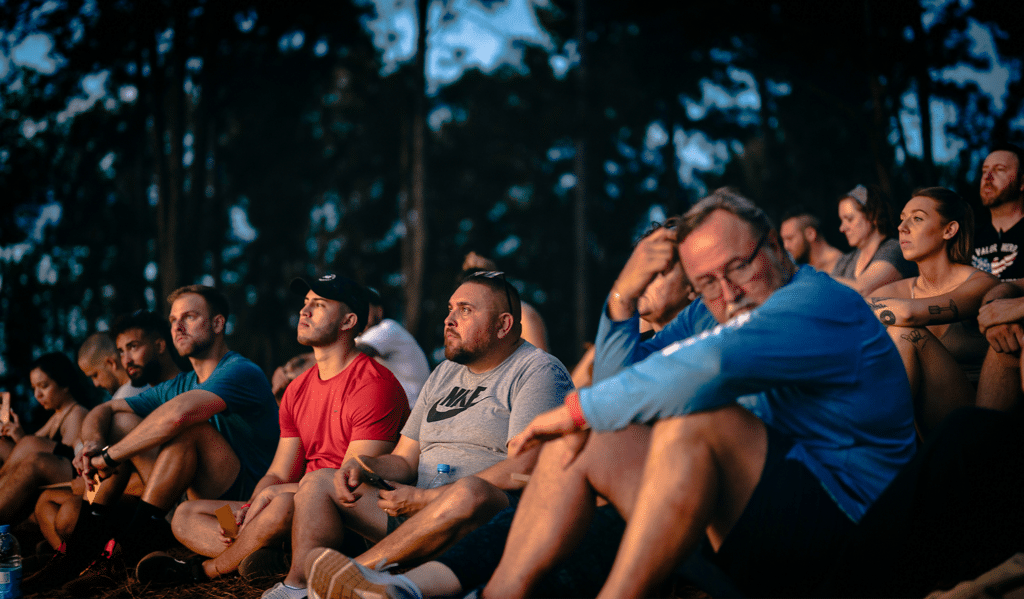 A group of people sitting outdoors in a wooded area during twilight. They appear focused and relaxed, with some holding drinks. The background is blurred with trees silhouetted against the evening sky.