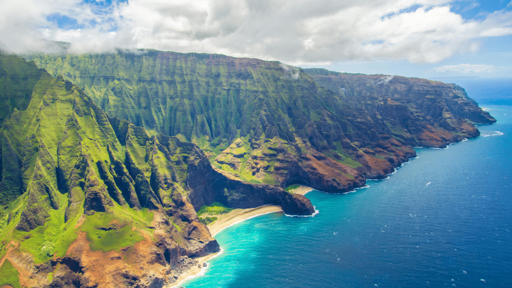 Aerial view of a rugged coastline with steep, green cliffs and a narrow sandy beach. The turquoise ocean waves crash against the shore, and the sky is partly cloudy, casting shadows on the landscape.