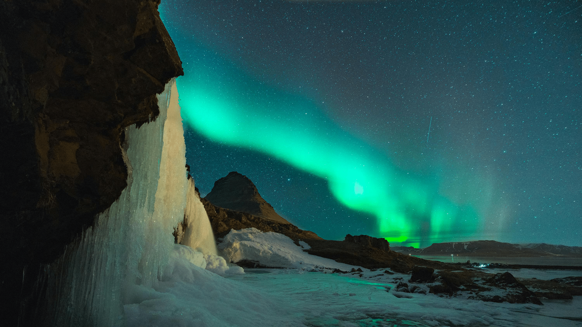 Aurora borealis lights up the night sky over a snowy landscape with a mountain in the background. A frozen waterfall is visible on the left, and stars twinkle across the clear sky.