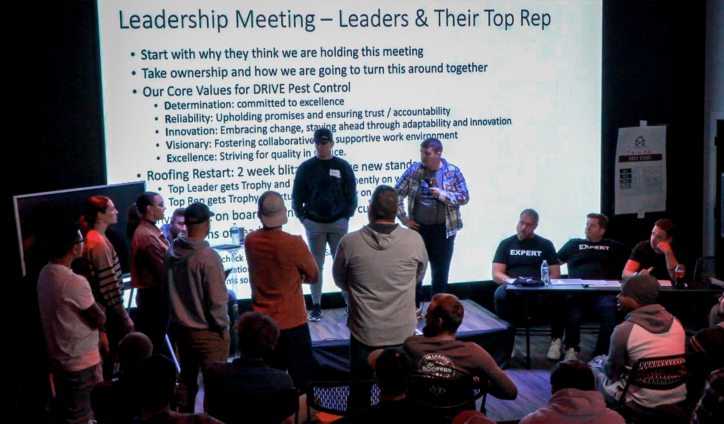 A group of people are gathered in front of a presentation screen displaying a leadership meeting agenda. The bullet points list core values for a pest control company. Some individuals are seated, while others stand at the front of the room.