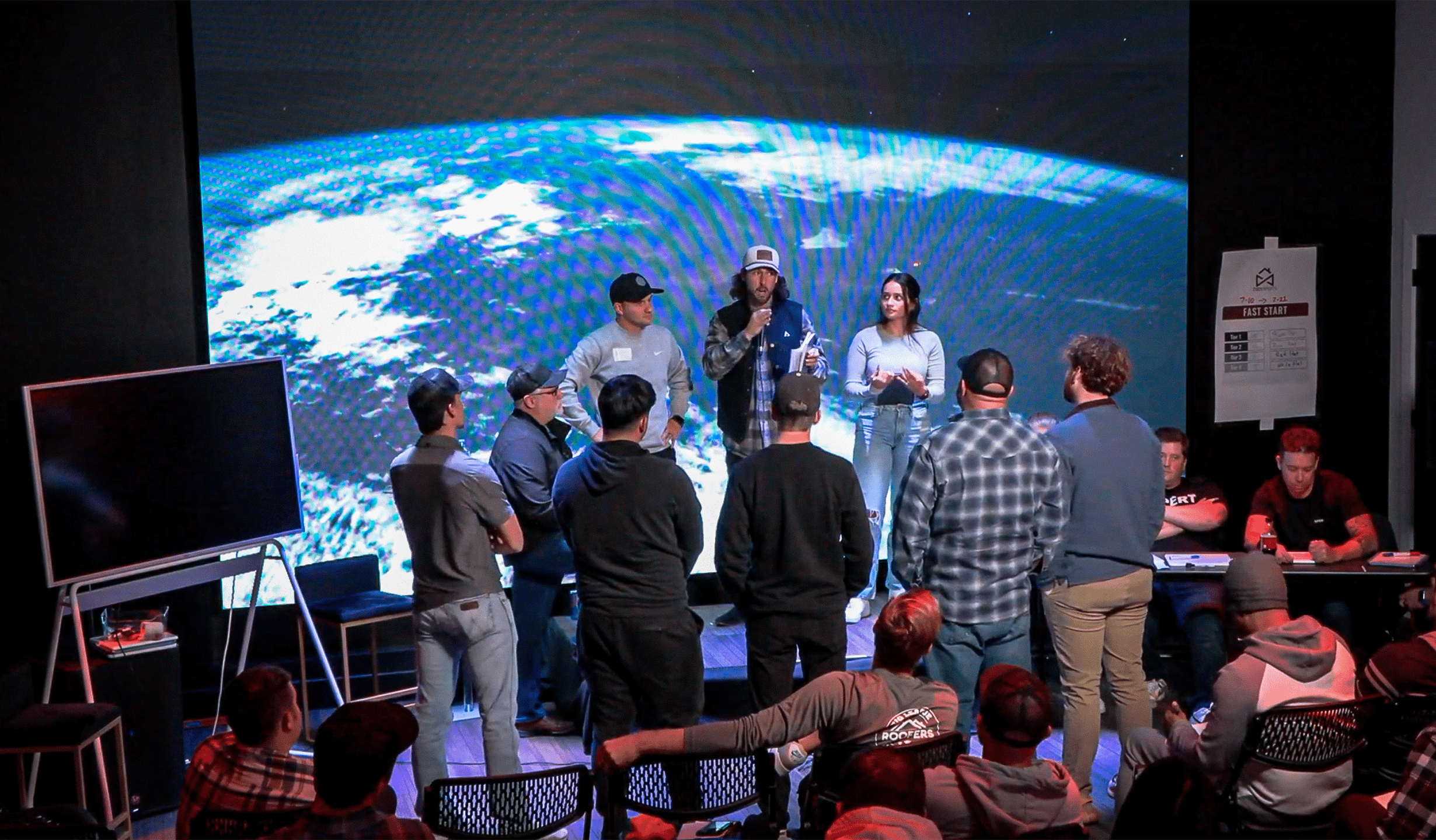 A group of people stands in a room with a large screen displaying an image of Earth from space. Some are seated, while others stand around a table. Two individuals hold microphones, possibly speaking to the audience.