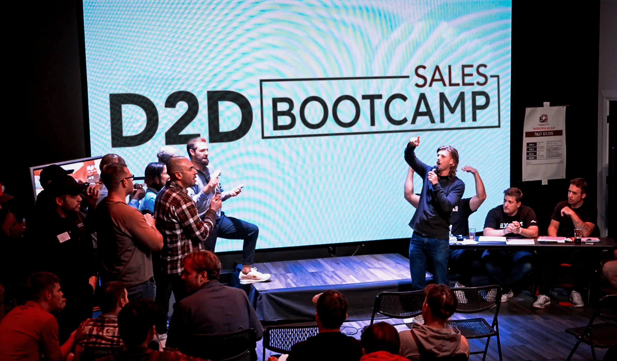 A lively speaker stands with one arm raised on stage at the "D2D Sales Bootcamp," as the audience claps and cheers. Several people are seated at a table with laptops on stage, while attendees watch from the seating area.