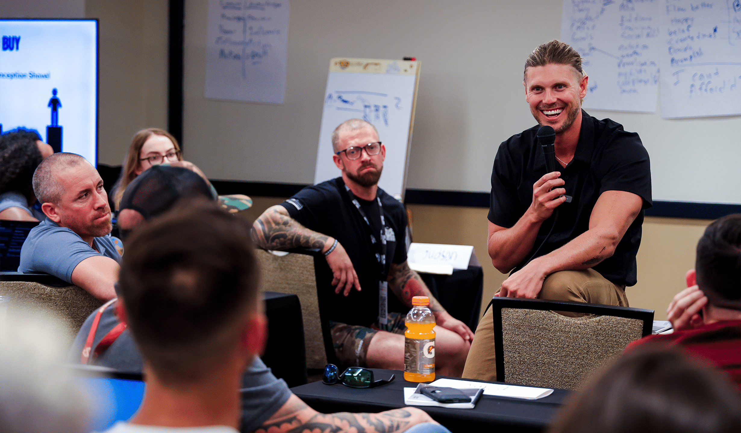 A man in a black shirt holds a microphone, speaking to a group in a classroom setting. Attendees sit attentively, with some taking notes. Charts and notes are visible on the walls. A bottle of orange drink is on a table in the foreground.