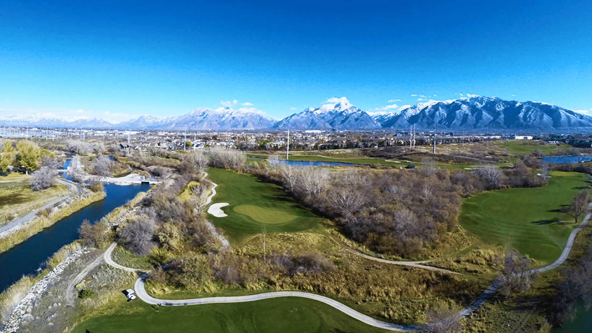 Aerial view of a golf course surrounded by trees and a winding path. A river flows through the landscape. Snow-capped mountains rise in the background under a clear blue sky.