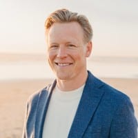 Smiling man with light hair wearing a blue blazer and white shirt stands on a sandy beach with the ocean and sky softly blurred in the background.