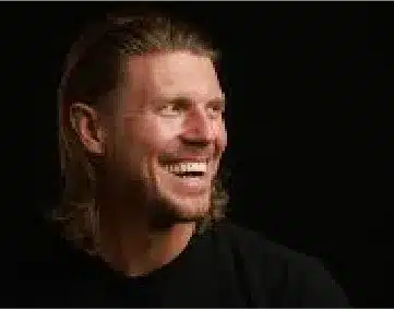 A man with long, light brown hair and a beard smiles broadly while looking to the side. He is wearing a black shirt and is set against a dark background.